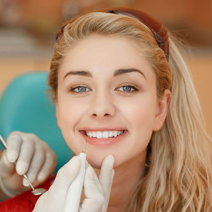 a young woman with blonde hair smiles at the camera while a gloved dentist hand holds a tool up to her teeth